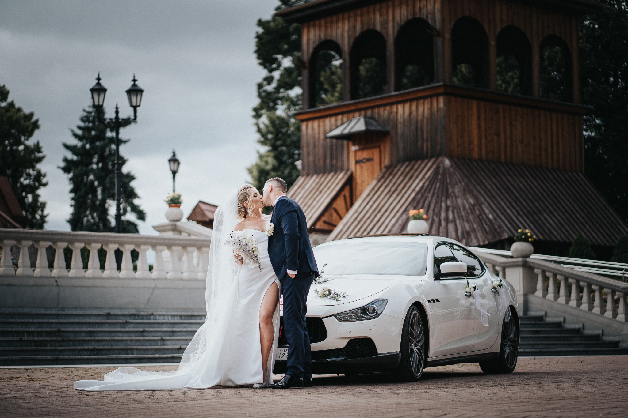 Pocałunek młodej pary w sukni ślubnej i garniturze na tle białego samochodu ozdobionego kwiatami i drewnianej architektury.