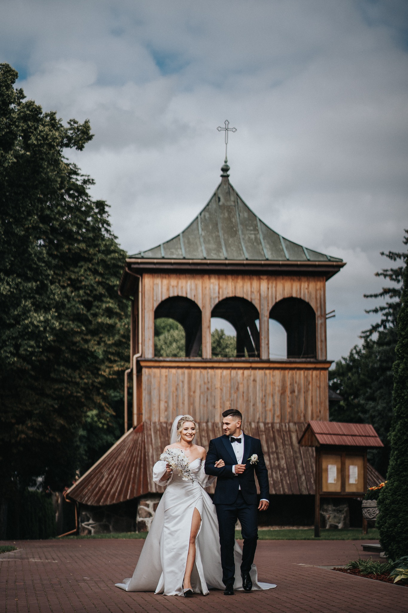 Para młoda spaceruje po ślubie na tle drewnianej wieży z krzyżem, panna młoda w sukni z rozcięciem i bukietem, pan młody w granatowym garniturze z muszką.
