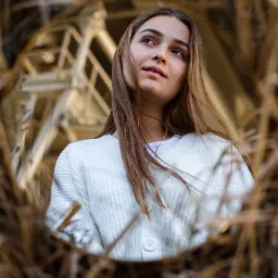 Portret młodej kobiety z długimi włosami, ubrana w biały sweter, patrzy w górę, sfotografowana przez ramę z suchych traw na tle metalowej konstrukcji.