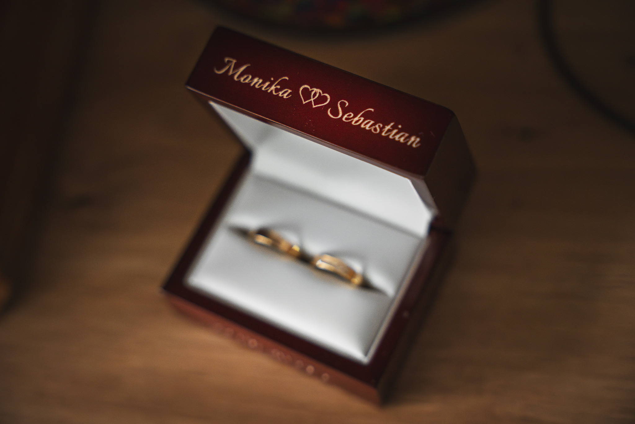 Otwarta, mahoniowa szkatułka z wygrawerowanymi imionami 'Monika' i 'Sebastian' oraz symbolami serc, prezentująca parę złotych obrączek na białej poduszeczce.