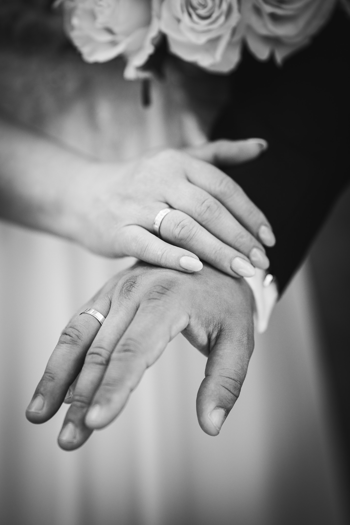 Czarno-białe zbliżenie dłoni pary z obrączkami na tle bukietu ślubnego. Delikatne światło podkreśla detale biżuterii i fakturę skóry.