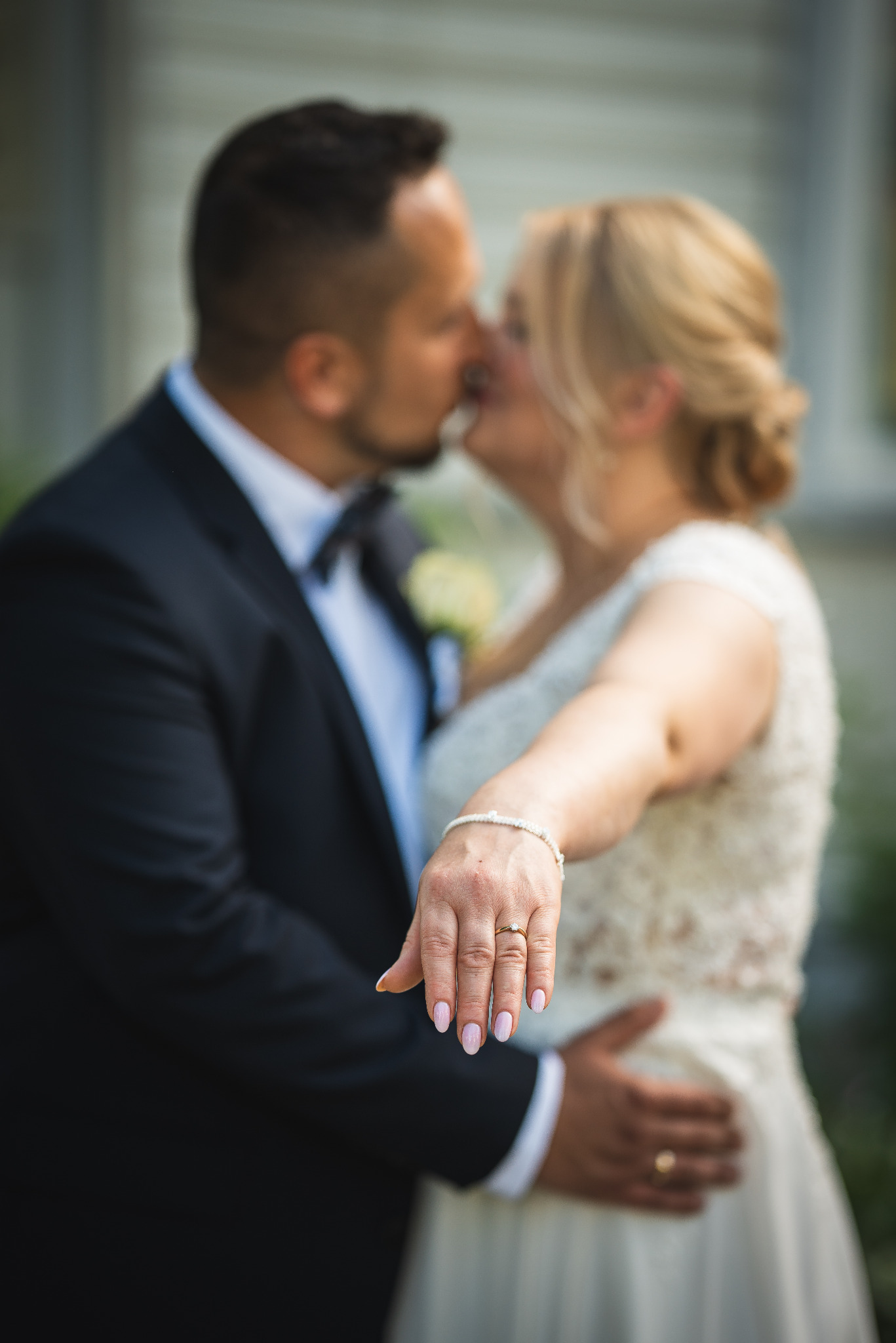 Pocałunek pary młodej w tle, z wyeksponowanym pierścionkiem zaręczynowym na dłoni panny młodej, skierowanej w stronę widza. Romantyczna atmosfera.