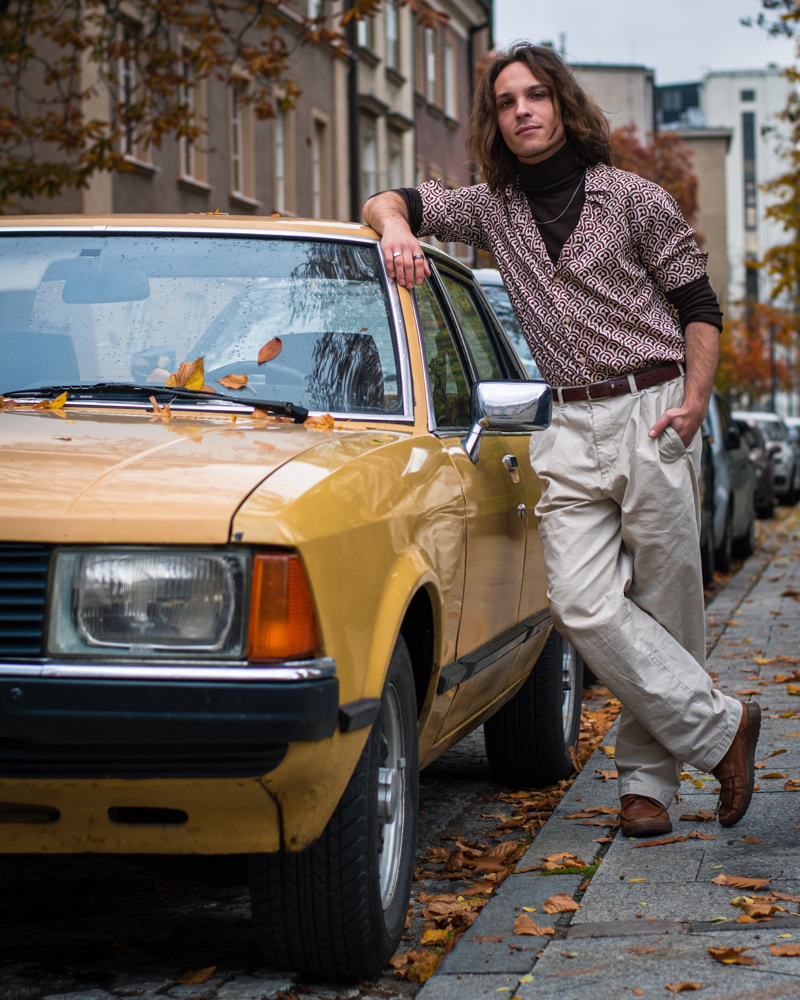 Mężczyzna w stylu retro opiera się o żółty samochód na tle miejskiej ulicy jesienią, liście na chodniku i na samochodzie.