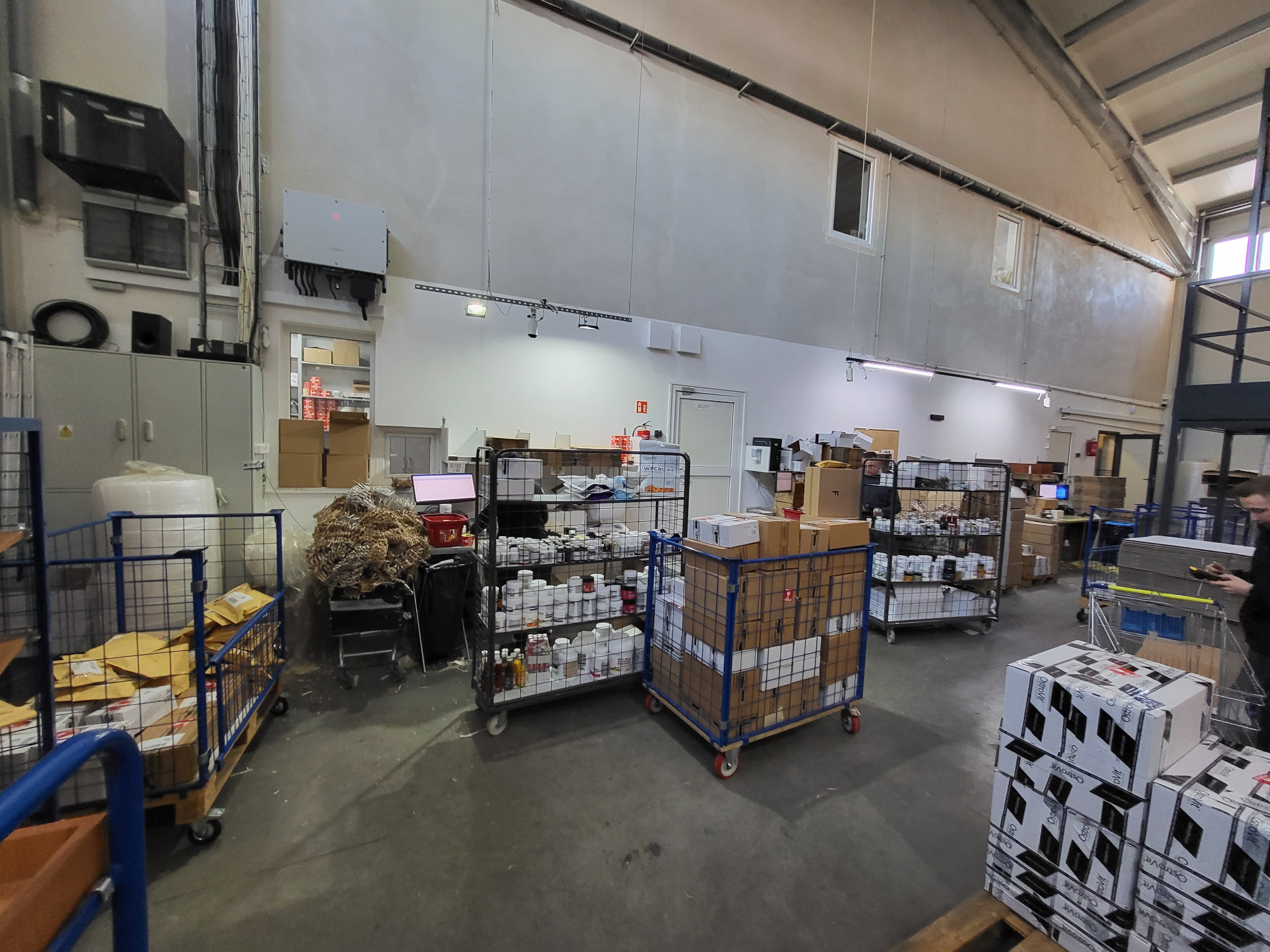 Wnętrze magazynu z metalowymi wózkami wypełnionymi paczkami, pudełkami i dokumentami, widoczne regały z zapasami, osoba w tle obsługująca skaner.