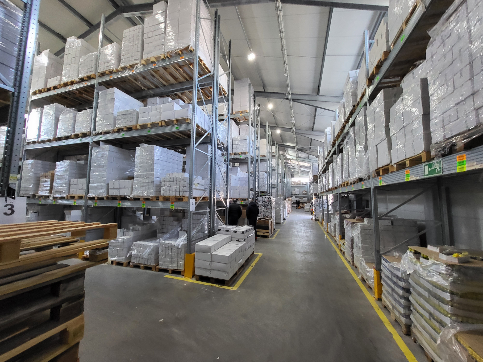 Wnętrze magazynu z wysokimi regałami wypełnionymi paletami z białymi, zapakowanymi produktami. Na podłodze widoczne żółte linie wyznaczające ścieżki. W tle dwie osoby w ciemnych ubraniach.