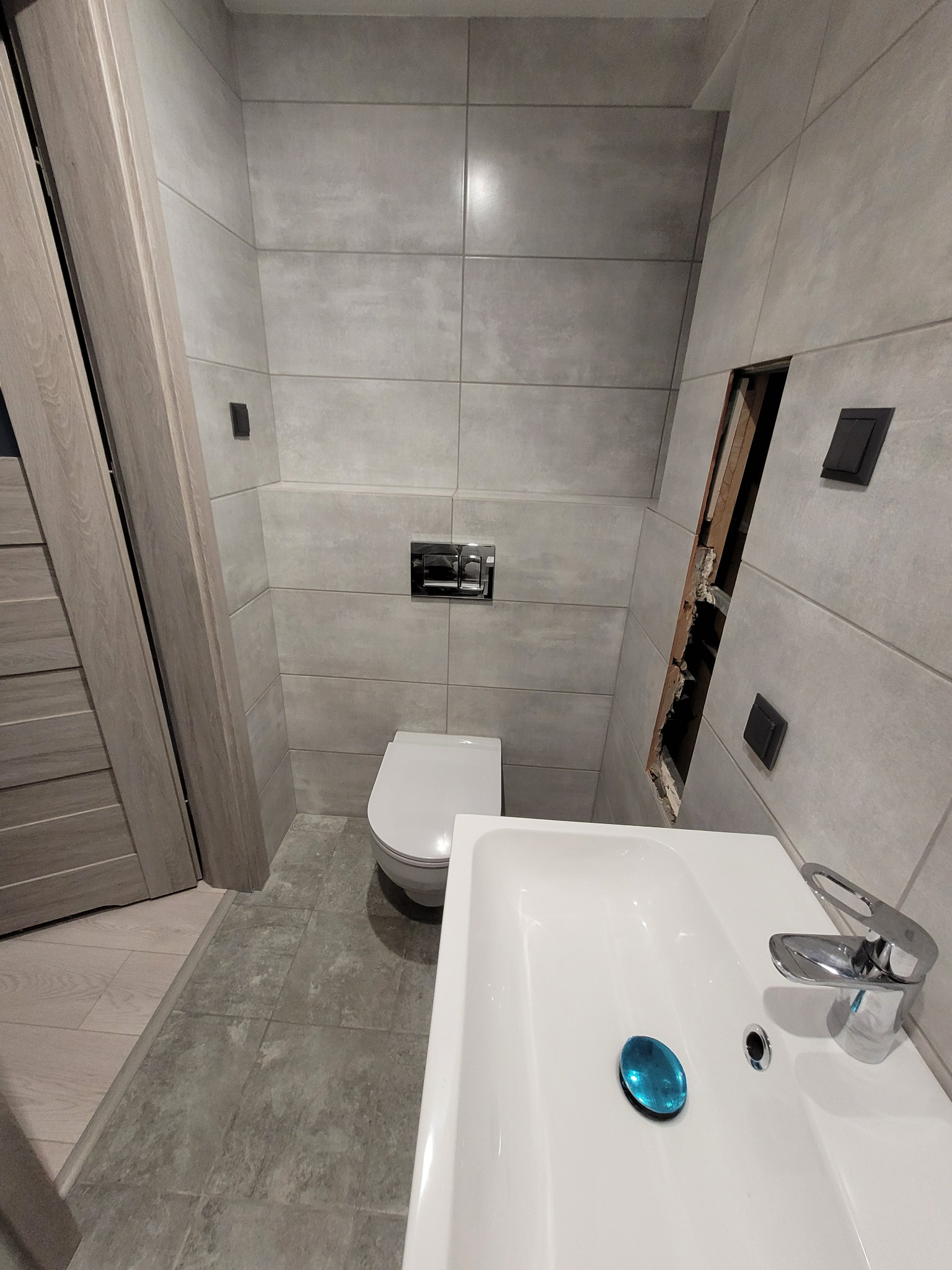 Wykończona łazienka z szarymi płytkami, białą umywalką z niebieską folią ochronną na odpływie, wiszącą toaletą i otwartym otworem w ścianie z widoczną konstrukcją.