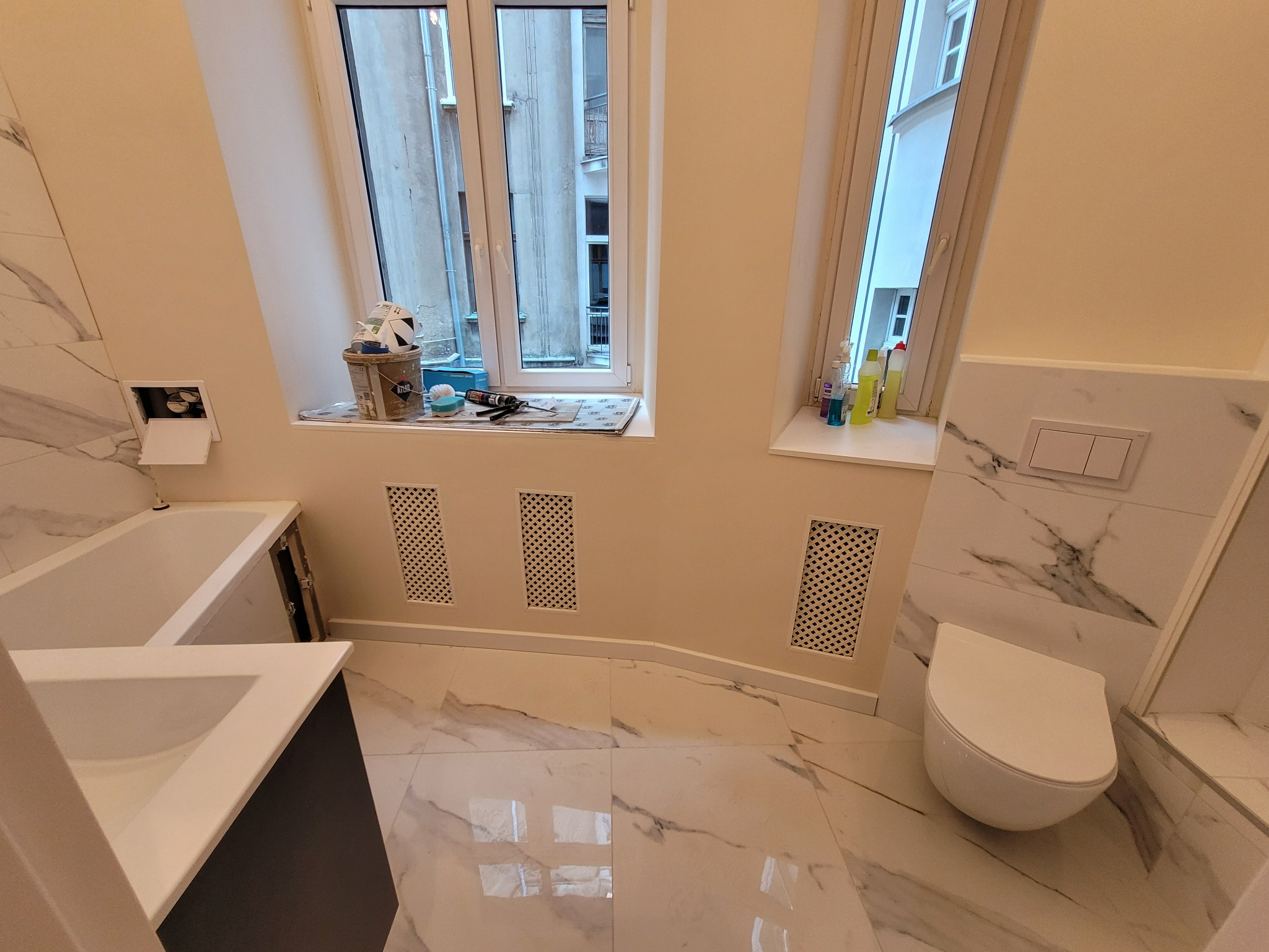 Wykończona łazienka z marmurowymi płytkami na ścianach i podłodze, widoczne okna z widokiem na budynki, biała toaleta i umywalka z szarą szafką, na parapecie okiennym stoją środki czystości...