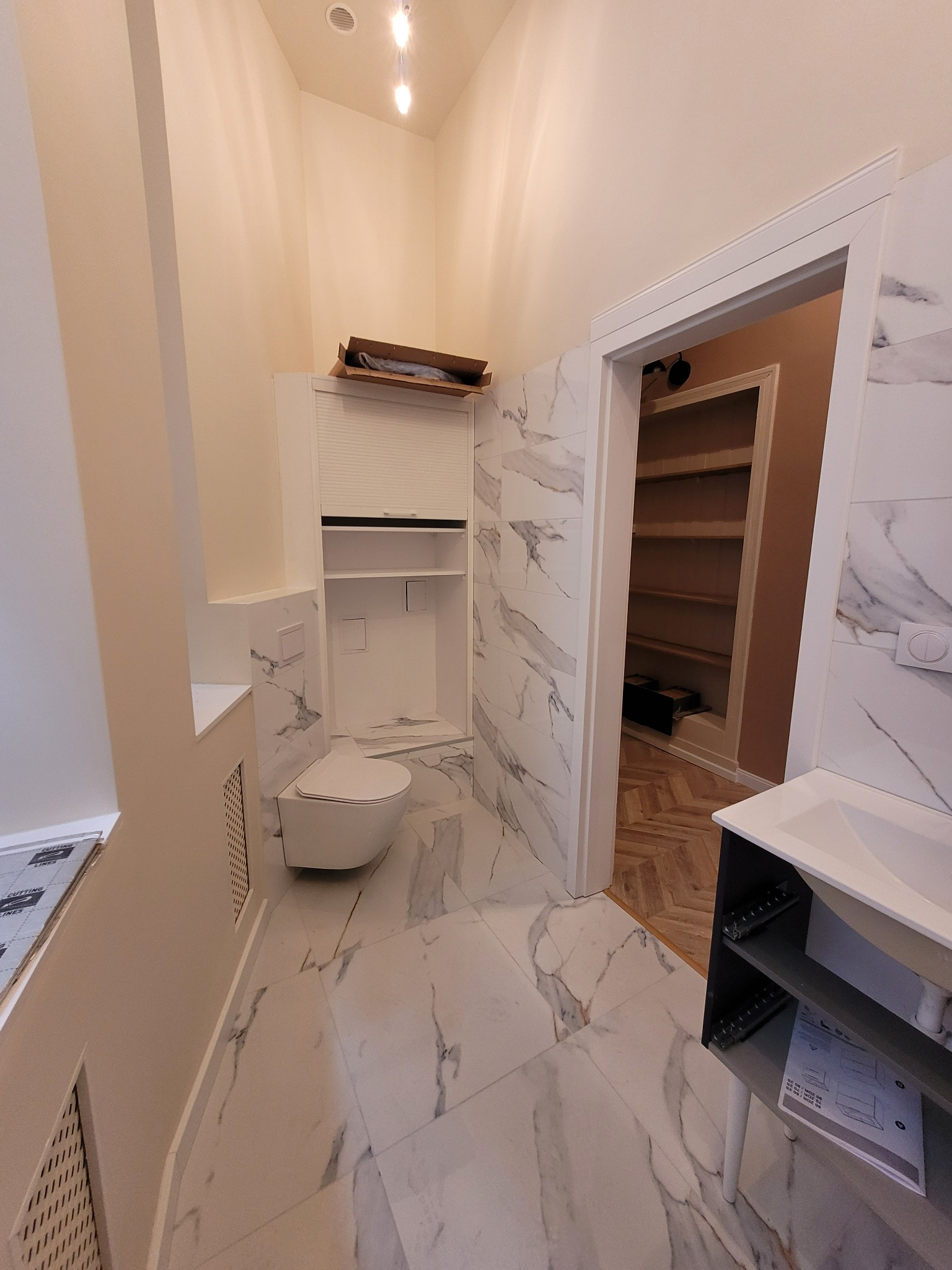 Nowoczesna łazienka z marmurowymi płytkami, podwieszaną toaletą, szafką z roletą i widokiem na garderobę z parkietem w jodełkę.