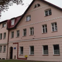 Dwupiętrowy budynek z różową elewacją, czerwonym dachem i białymi oknami, z szyldem sklepu z odzieżą i pasmanterią na parterze.