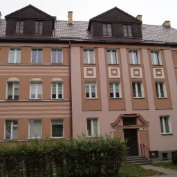 Wielorodzinny budynek z ocieploną elewacją w odcieniach beżu i brązu, z dekoracyjnymi elementami architektonicznymi nad oknami i ciemnym dachem z lukarnami.