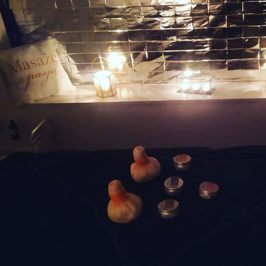 Przytulne wnętrze gabinetu masażu z poduszką z napisem 'Masaże z pasją', świecami w lampionach, srebrną ścianą w tle oraz dwoma bawełnianymi woreczkami z ziołami i zestawem małych słoiczków...