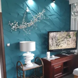 Turkusowa ściana z trójwymiarowymi panelami, oświetlona punktowo, z dekoracją z gałązek, lampa stołowa z ceramiczną podstawą na wiklinowym stoliku, telewizor na szafce RTV z ciemnego drewna.