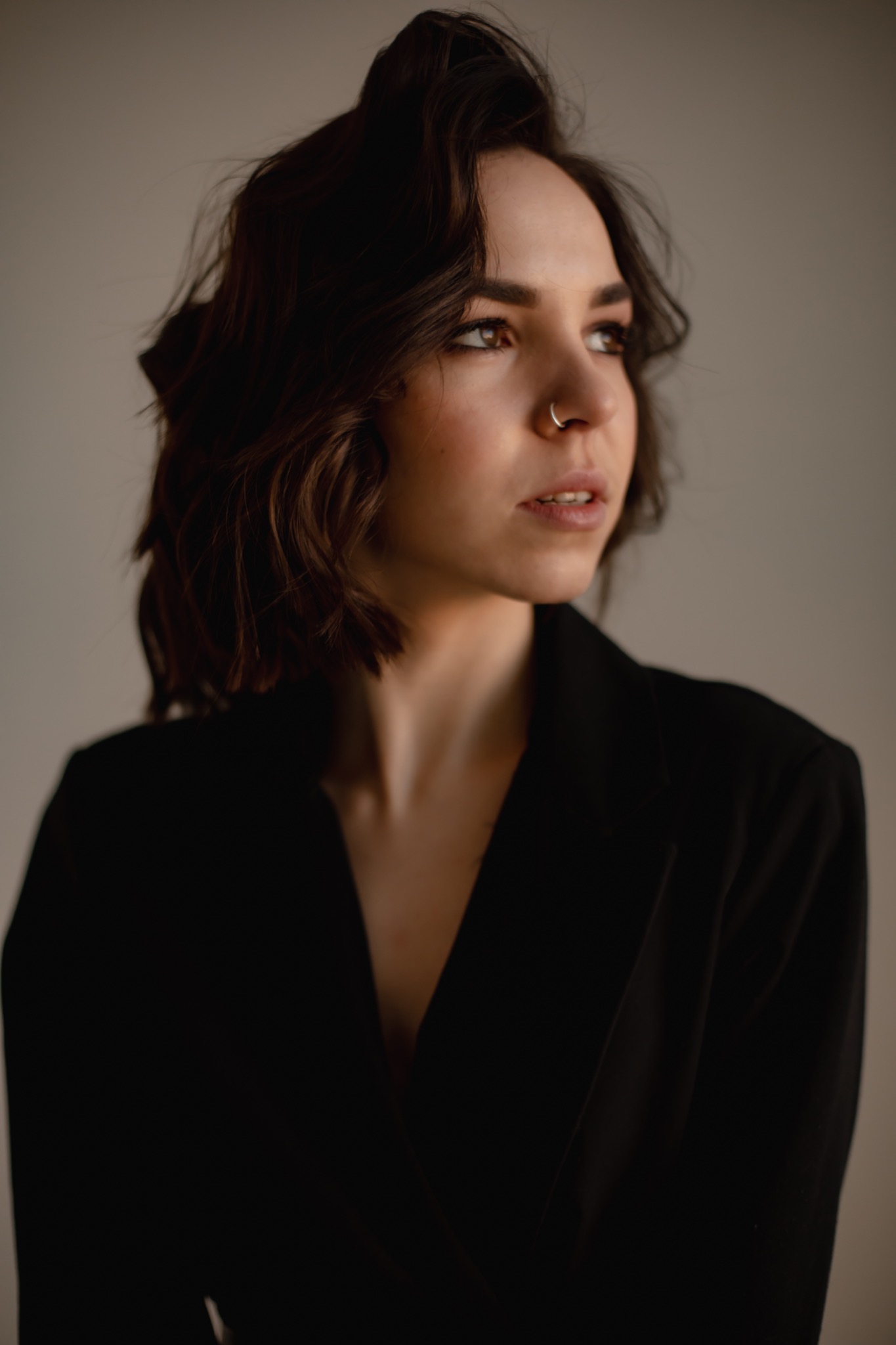 Portret kobiety w czarnej marynarce z kolczykiem w nosie, spoglądającej w bok na neutralnym tle.