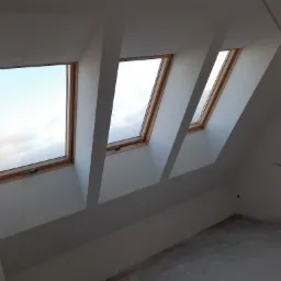 Trzy okna dachowe z drewnianymi ramami w nowo wykończonym poddaszu z gładkimi, białymi ścianami i surową podłogą. Wisząca żarówka oświetla wnętrze.