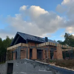 Budowa domu w stanie surowym otwartym: widoczne mury z czerwonej cegły, szalunki stropowe z desek, fundamenty z szarego betonu komórkowego, czarny dach i błękitne niebo z chmurami w tle.