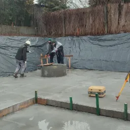 Prace na budowie: dwóch mężczyzn w odzieży roboczej analizuje dokumenty obok betonowego kręgu na świeżo wylanej płycie fundamentowej, w tle czarna folia zabezpieczająca skarpę i żółty niwelator...