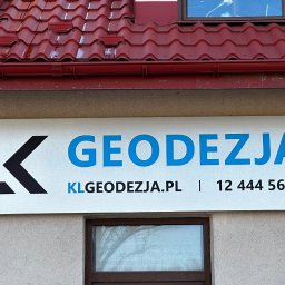 GEODEZJA S.C. Klimasara, Liwora - Szyld firmy z niebieskim napisem 'Geodezja' i czarnym logo 'K', umieszczony na elewacji budynku z czerwonym dachem i oknami. Widoczny numer telefonu i adres strony internetowej.