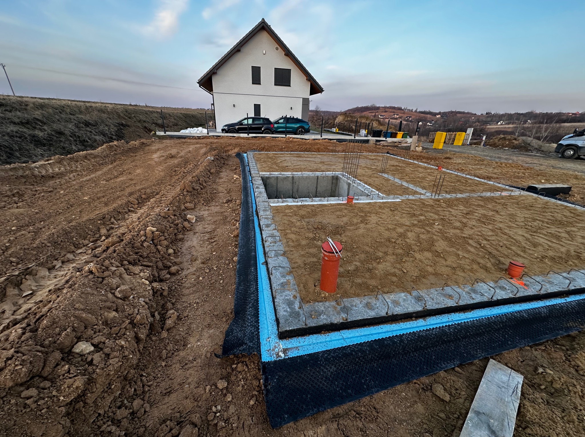 Fundamenty domu jednorodzinnego w trakcie budowy, widoczne zbrojenia i izolacja, w tle budynek w stanie surowym otwartym i dwa samochody osobowe.