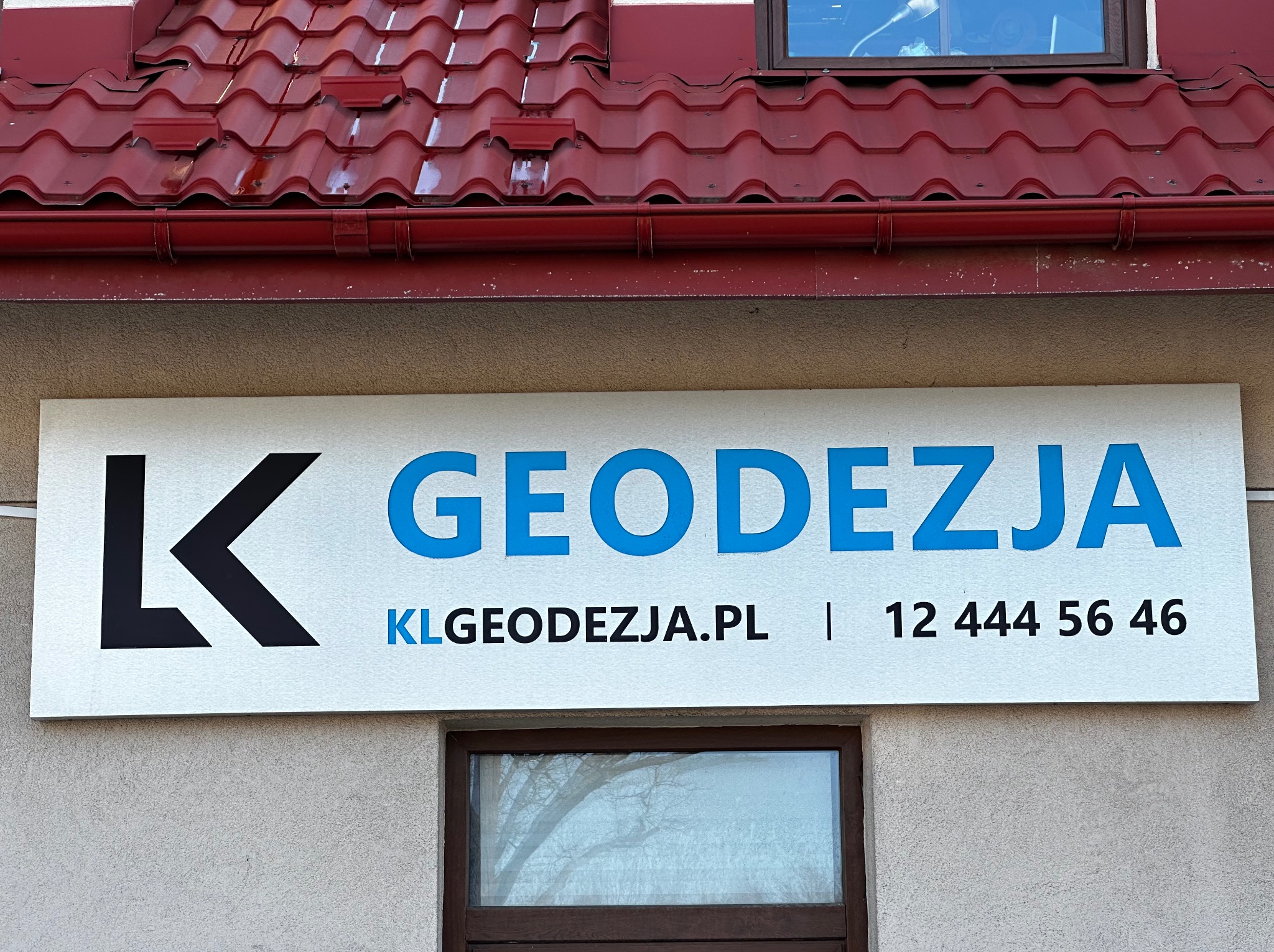 Szyld firmy z niebieskim napisem 'Geodezja' i czarnym logo 'K', umieszczony na elewacji budynku z czerwonym dachem i oknami. Widoczny numer telefonu i adres strony internetowej.
