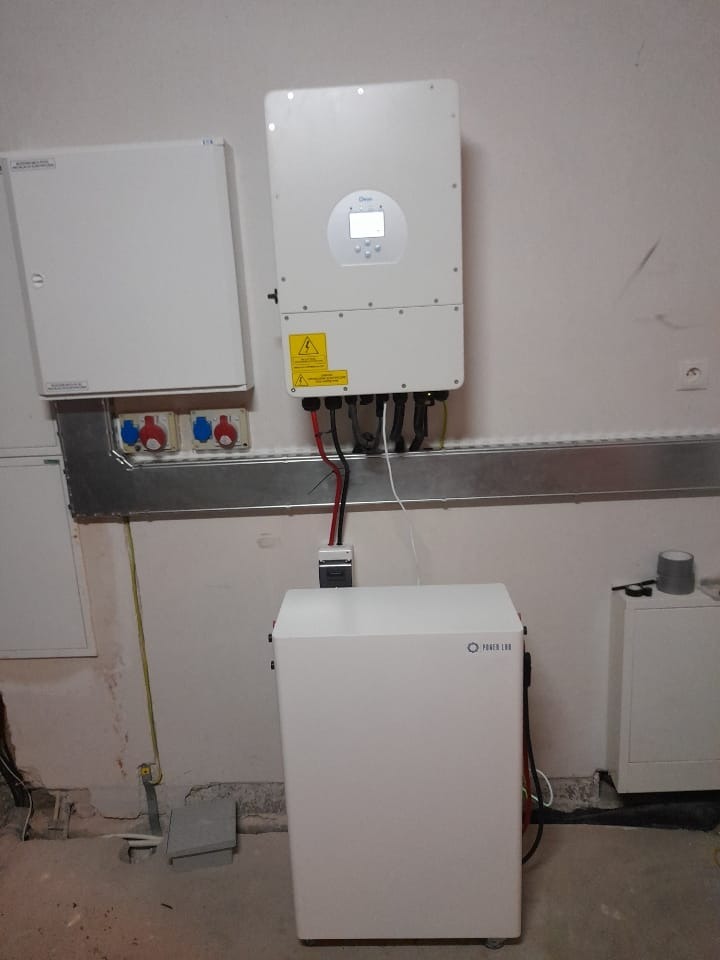 Biały akumulator energii z logo Power Lab podłączony do białego inwertera na ścianie z widocznymi kablami i osprzętem elektrycznym.