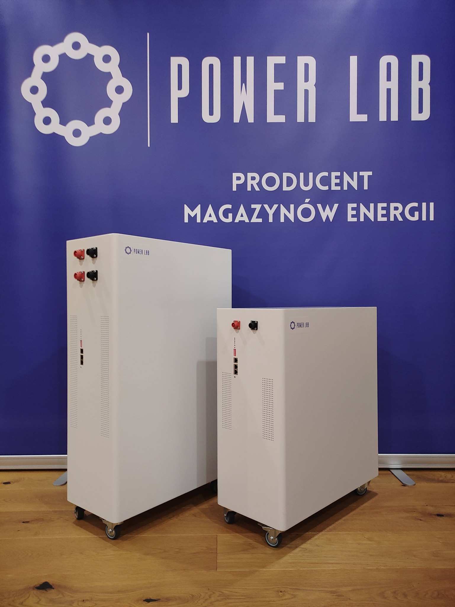 Dwa białe mobilne moduły zasilania na tle niebieskiego baneru z napisem 'Power Lab Producent Magazynów Energii', stojące na drewnianej podłodze.