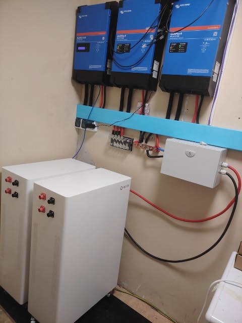 Instalacja magazynu energii: Dwa białe moduły bateryjne na kółkach, podłączone do trzech niebieskich inwerterów Victron Energy zamontowanych na ścianie z niebieską listwą. Widoczne okablowanie...