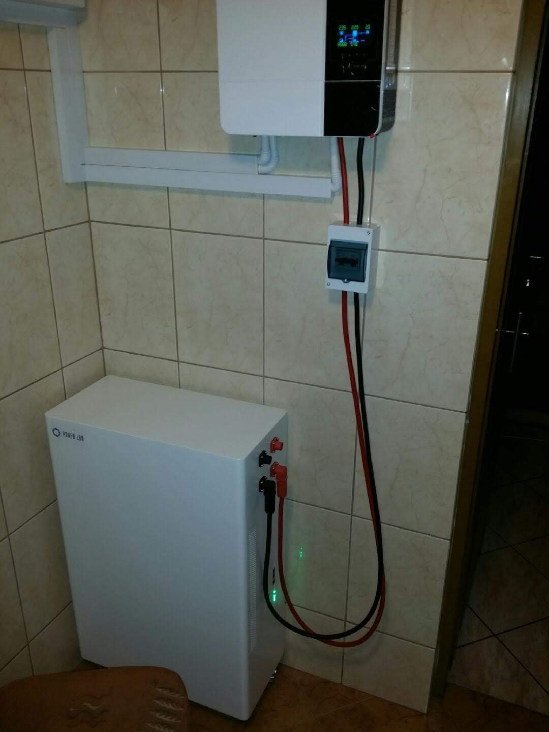 Biały akumulator energii Power Lab z czerwonymi i czarnymi kablami podłączony do białego inwertera na kafelkowanej ścianie. Widoczny wyłącznik bezpieczeństwa i fragment brązowego krzesła.