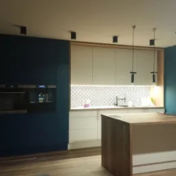 Nowoczesna kuchnia z granatowymi i białymi szafkami, wyspą z drewnianym blatem, oświetleniem punktowym i dekoracyjną mozaiką nad blatem.