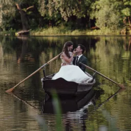 Para młoda siedząca w łodzi na spokojnym jeziorze, panna młoda dotyka nosa pana młodego, wiosła po bokach, drzewa w tle.