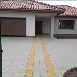 STEN-BUD Usługi Brukarskie - Nowoczesny podjazd z jasnoszarej kostki brukowej z dwoma pasami żółtej kostki prowadzącymi do domu z brązową bramą garażową i otwartą, zieloną furtką.