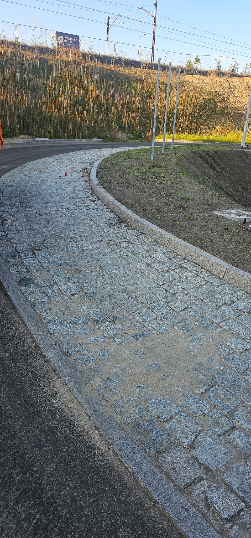 Zakrzywiona ścieżka wyłożona szarą kostką brukową, obrzeża z szarego granitu, widoczna ziemia i trawa po prawej stronie, asfalt po lewej, w tle wzgórze z trawą i słupami trakcji elektrycznej.