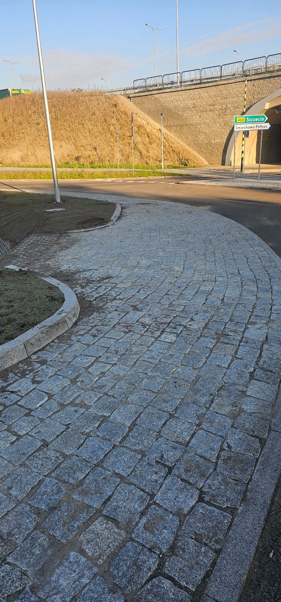 Ułożona z kostki granitowej ścieżka o nieregularnym kształcie, obramowana betonowym krawężnikiem, prowadząca w kierunku drogi i tunelu pod nasypem.