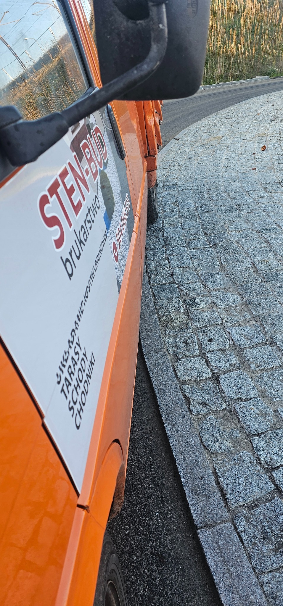Pomarańczowy samochód dostawczy firmy brukarskiej zaparkowany obok nowo ułożonej kostki brukowej z szarej kostki granitowej, widoczne logo firmy 'STEN-BUD' i numer telefonu.