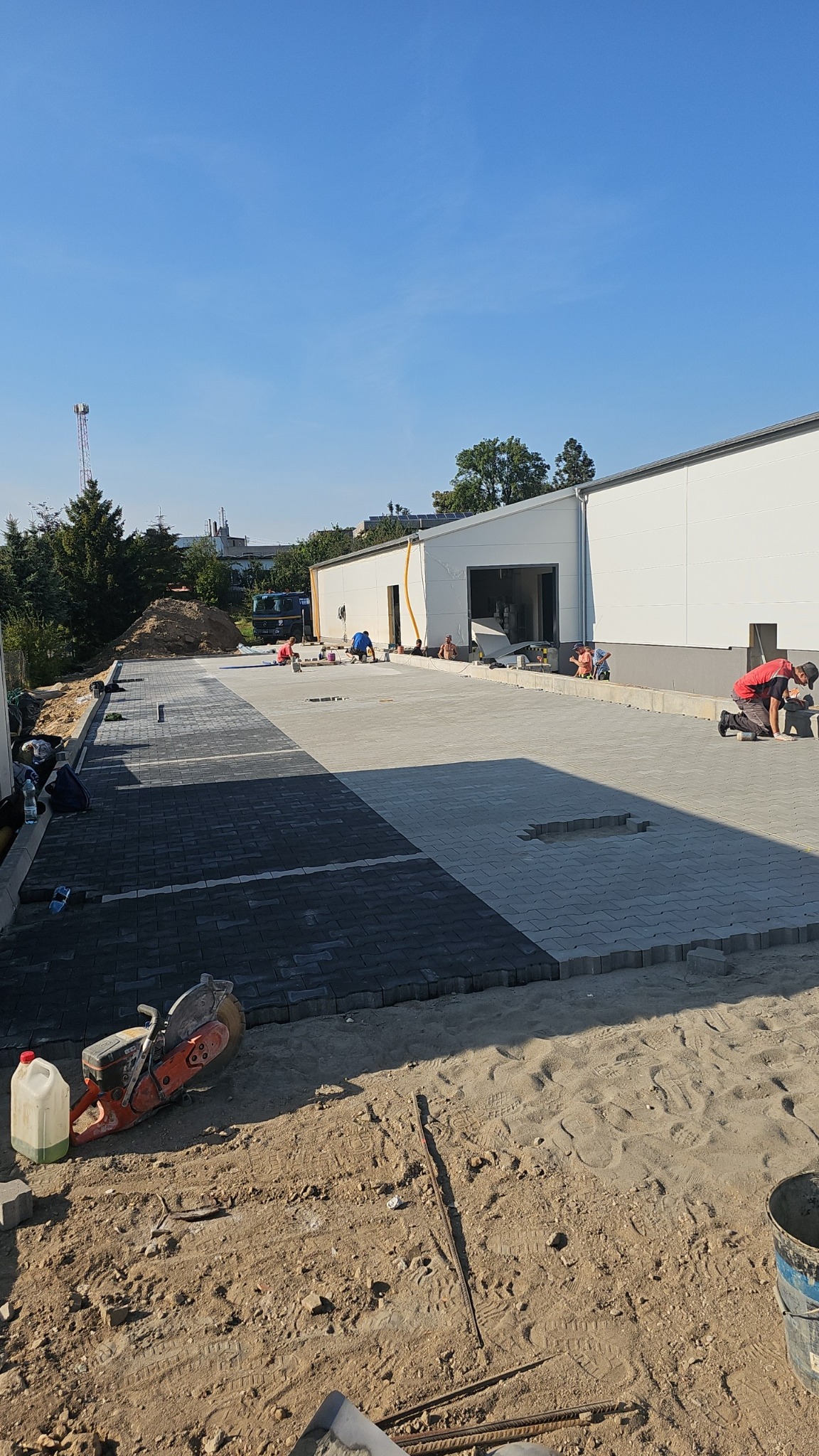 Trwa układanie szarej i grafitowej kostki brukowej na dużej powierzchni przed budynkiem magazynowym; widoczni pracownicy, piła do cięcia kostki, oraz częściowo ułożona nawierzchnia.