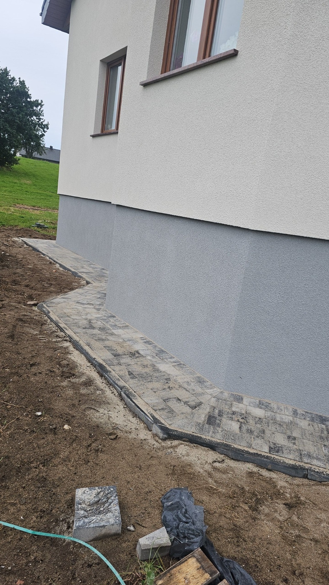 Ścieżka z szarej kostki brukowej ułożona wzdłuż ściany domu, z widocznymi elementami wykończeniowymi i pozostałościami po pracy w postaci ziemi i pojedynczych kostek.