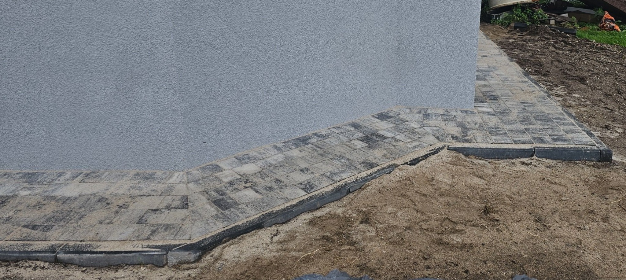 Ułożona kostka brukowa wzdłuż ściany budynku, zasypana piaskiem, widoczne krawężniki, ziemia i roślinność w tle.