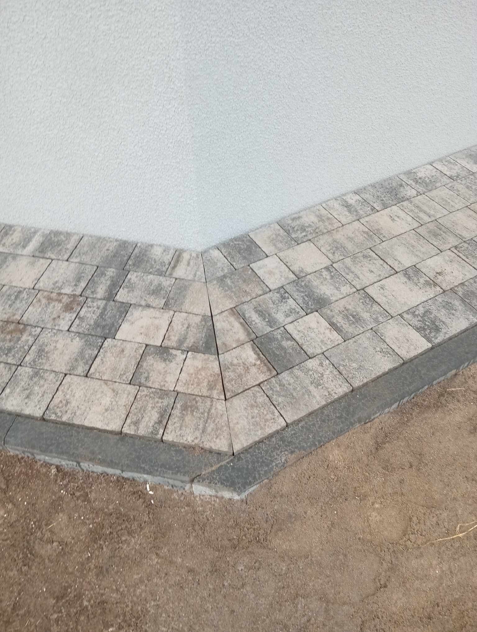 Ułożona kostka brukowa w odcieniach szarości i beżu, tworząca ścieżkę przy ścianie budynku z szorstkim tynkiem.