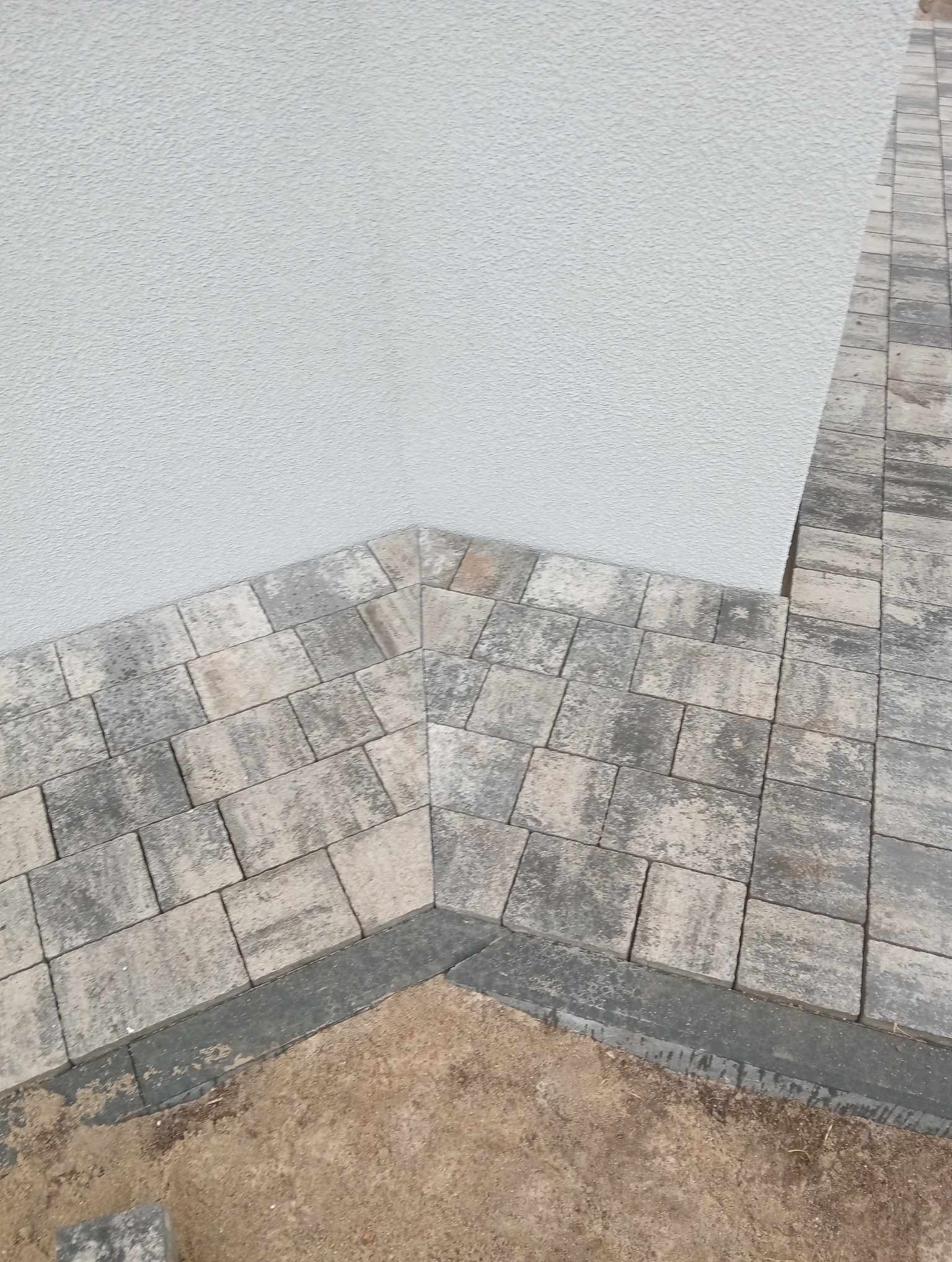 Szare i beżowe kostki brukowe ułożone przy białej ścianie budynku, widoczny fragment podsypki piaskowej.