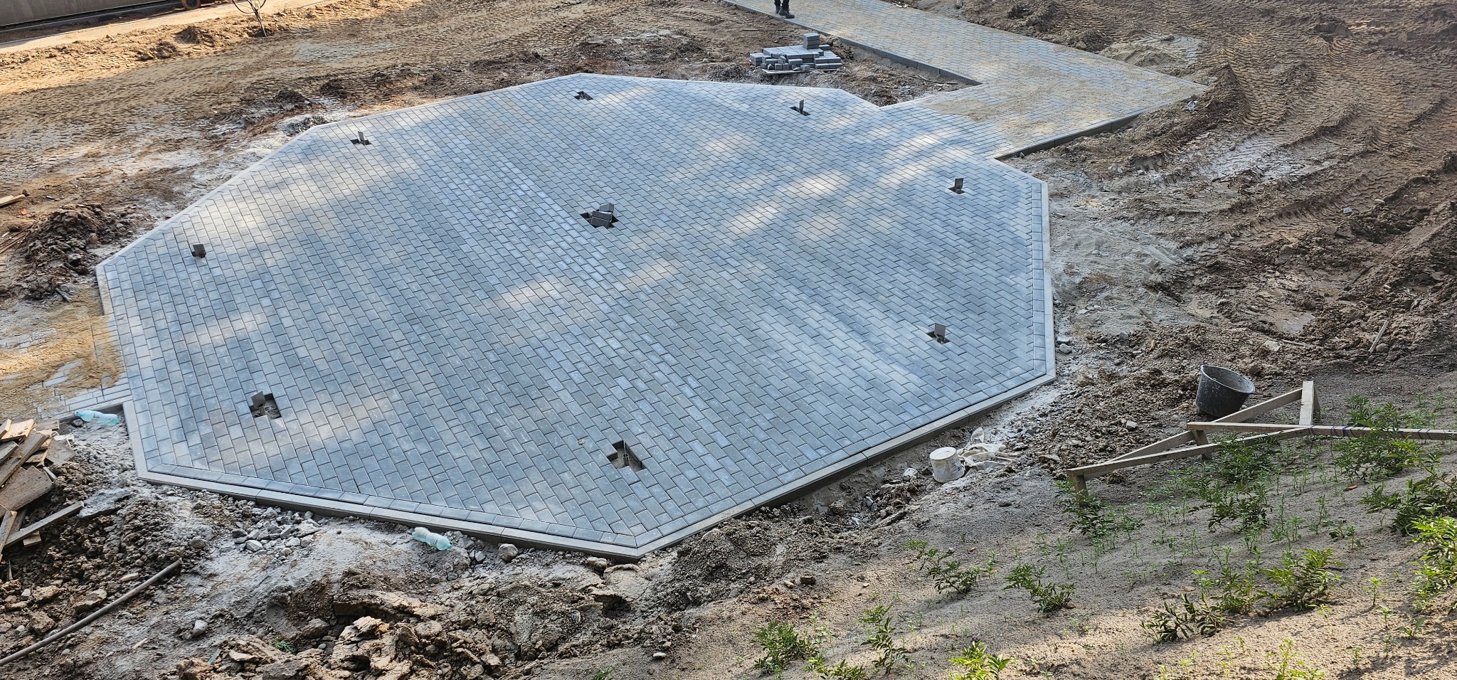 Niedokończona, ośmiokątna powierzchnia z szarej kostki brukowej z widocznymi otworami i brakującymi kostkami, otoczona ziemią i fragmentami ułożonego chodnika w tle.