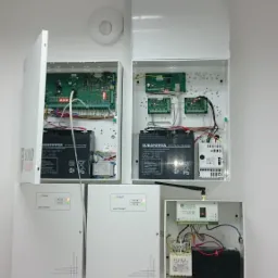 Rozbudowana instalacja zabezpieczeń z centralami alarmowymi, akumulatorami i modułami komunikacyjnymi w białych obudowach na ścianie. Widoczne okablowanie, płytki drukowane i sygnalizacja LED.