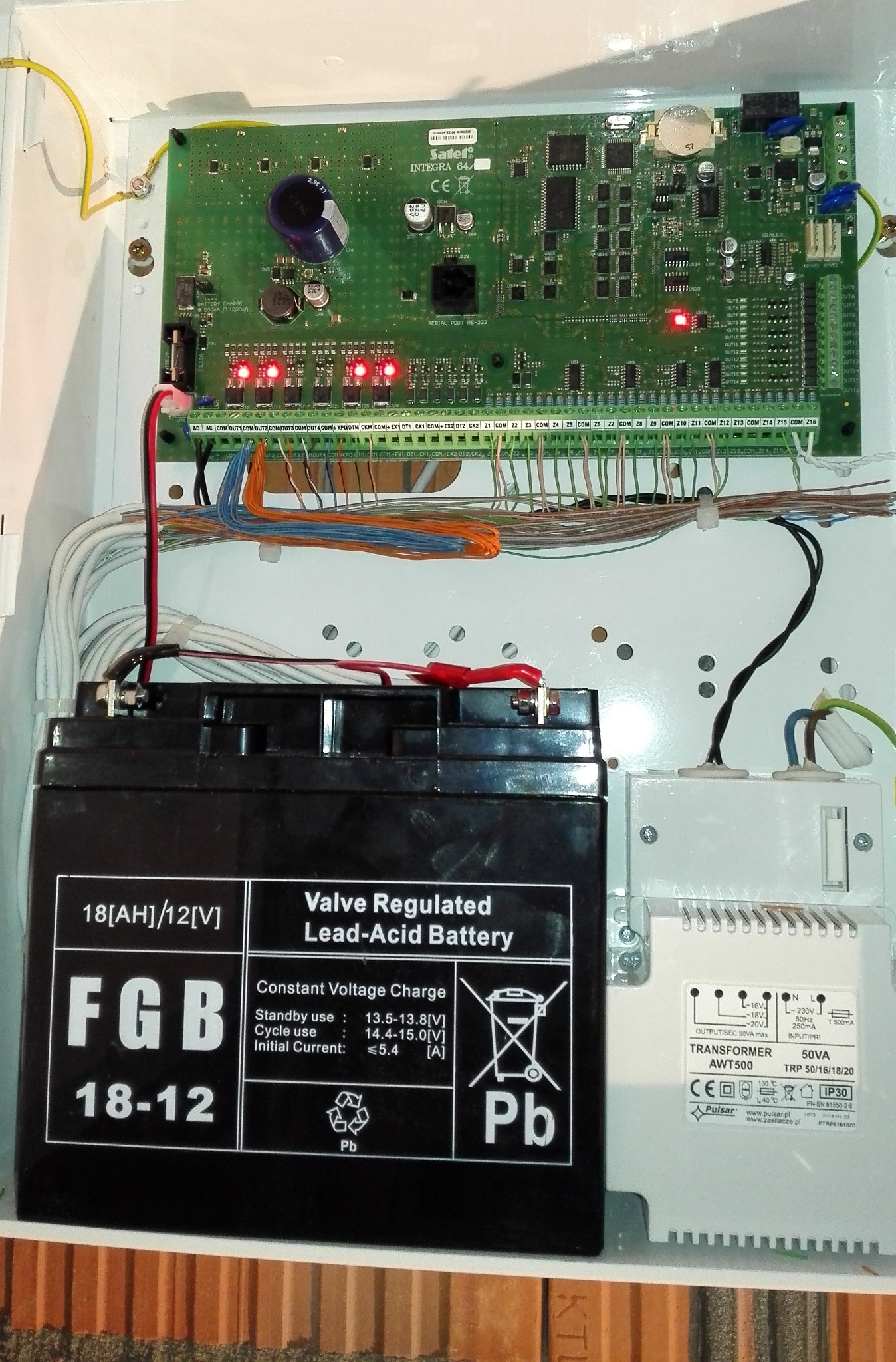 Otwarta obudowa centrali alarmowej z widoczną płytą główną Satel Integra 64, akumulatorem FGB 18-12 oraz transformatorem Pulsar AWT500, liczne przewody podłączone do listew zaciskowych.