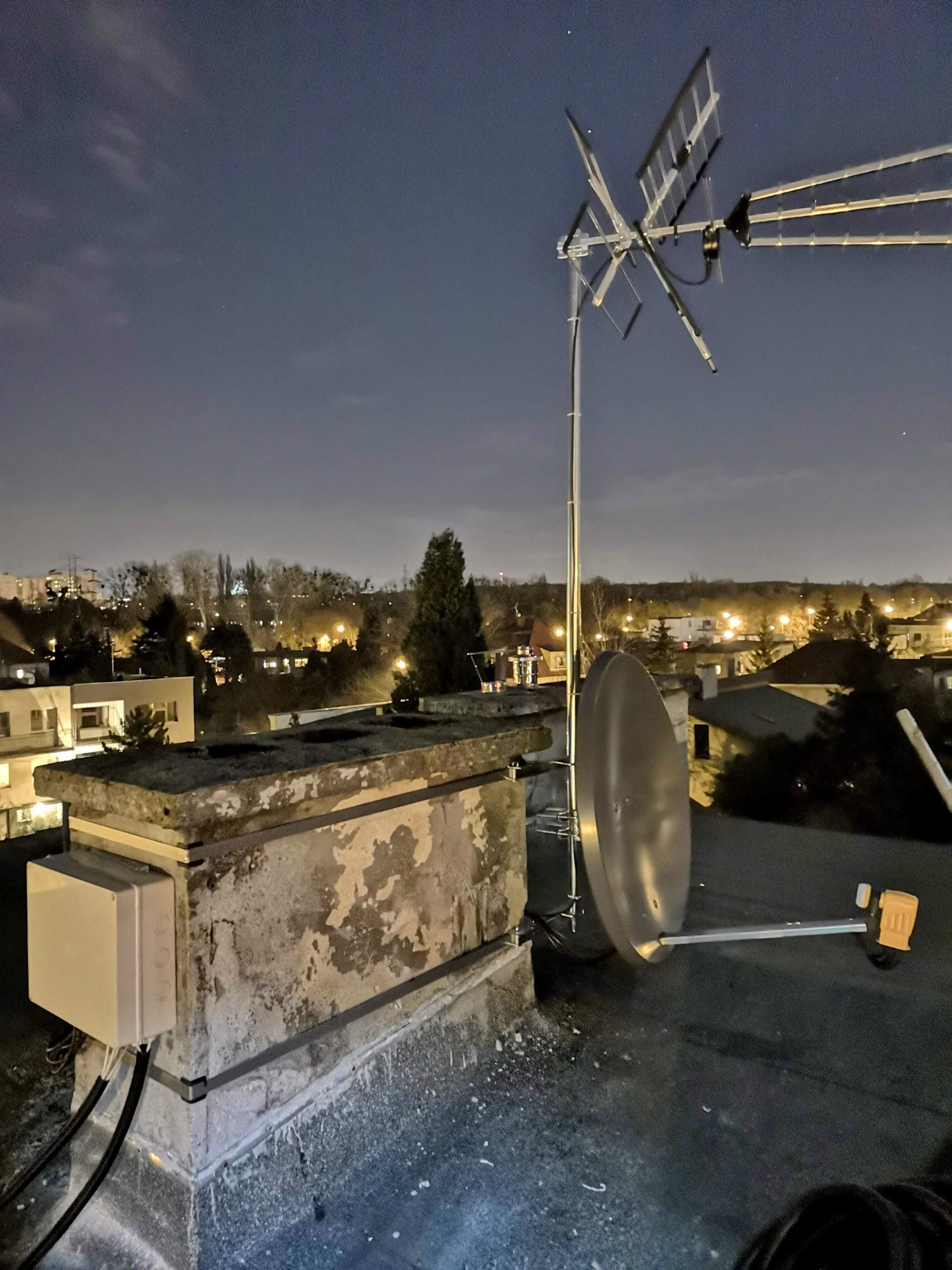 Antena satelitarna i antena telewizyjna zamontowane na dachu budynku w nocy, widoczny oświetlony krajobraz miejski w tle.