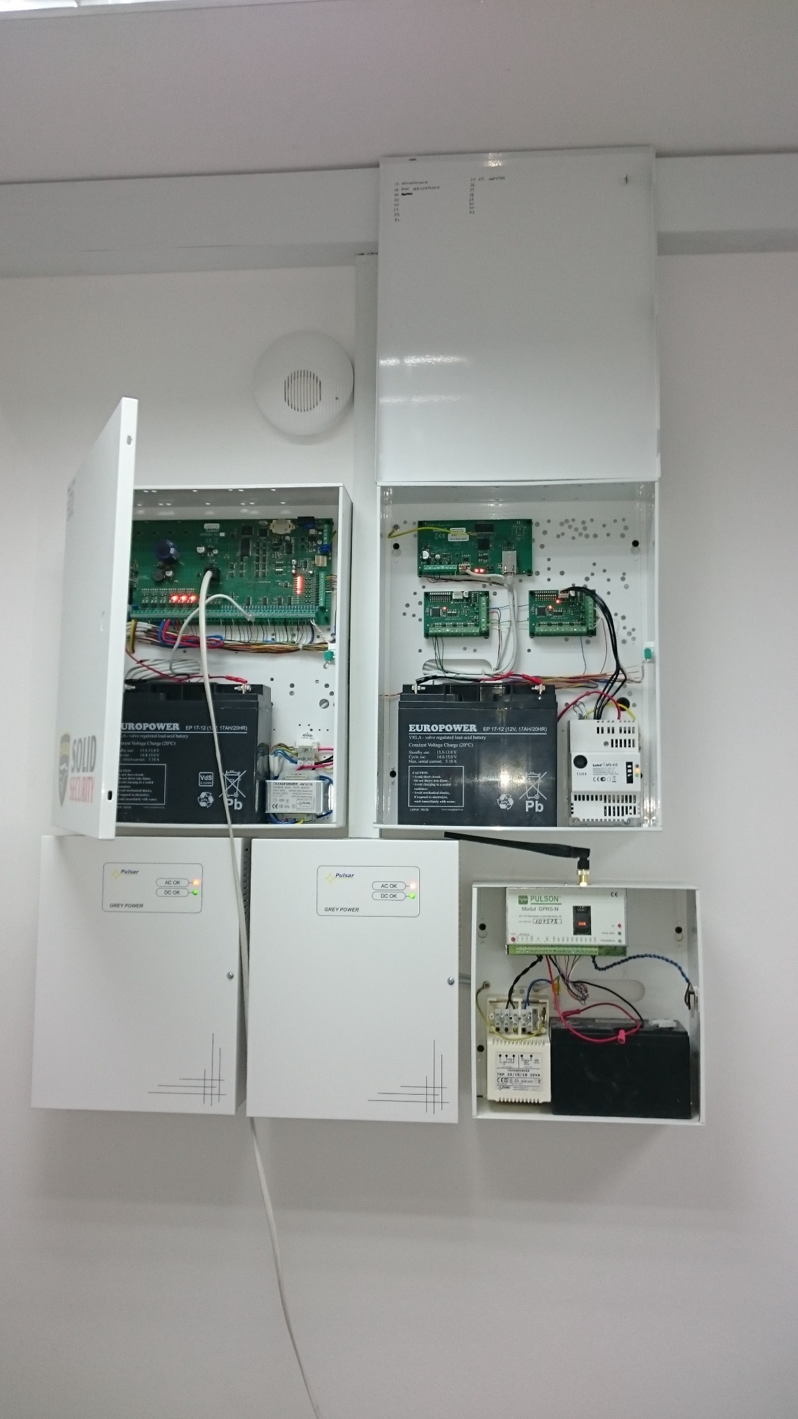 Rozbudowana instalacja zabezpieczeń z centralami alarmowymi, akumulatorami i modułami komunikacyjnymi w białych obudowach na ścianie. Widoczne okablowanie, płytki drukowane i sygnalizacja LED.