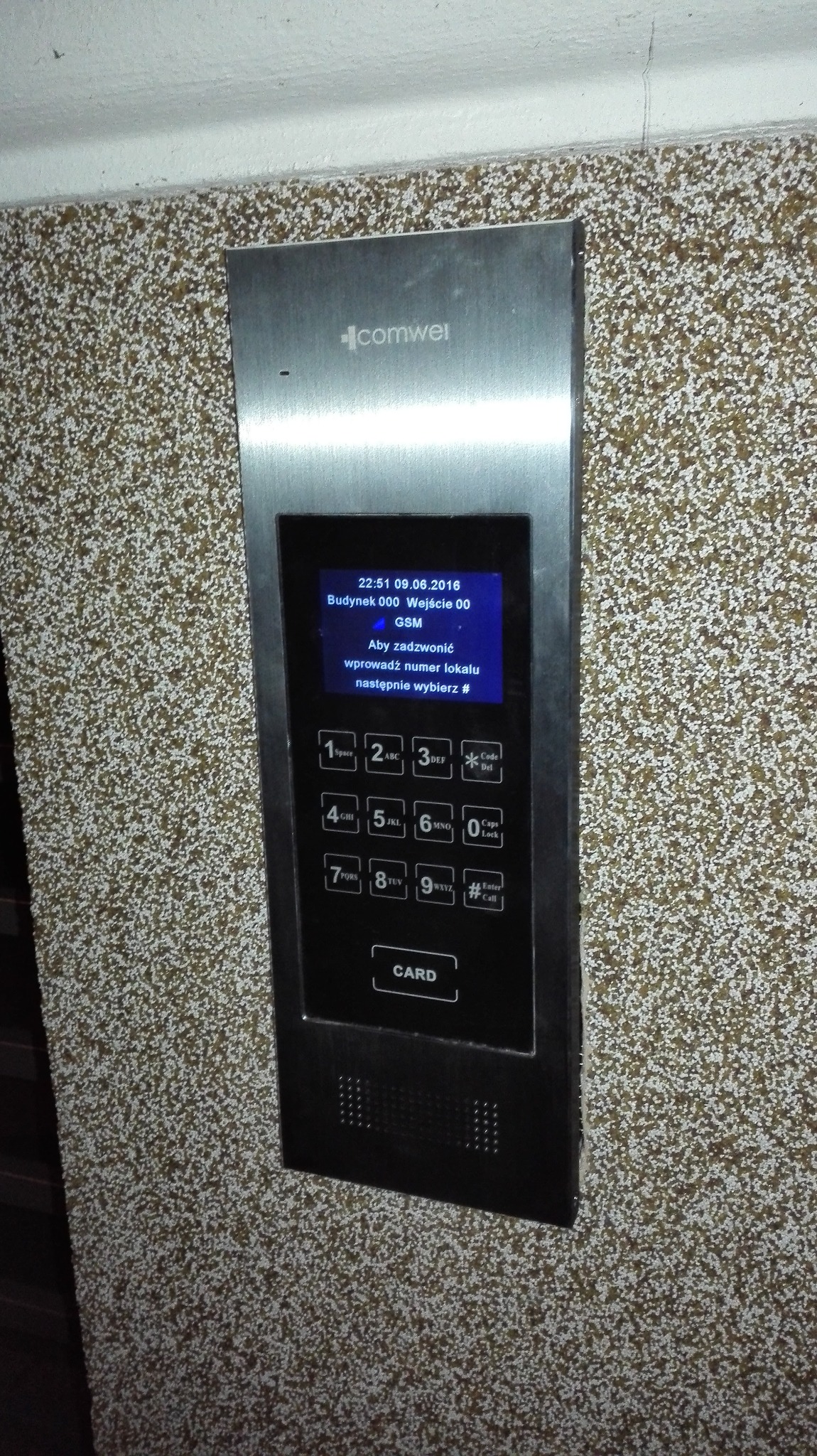 Panel domofonu cyfrowego Comwei z czytnikiem kart zbliżeniowych, zamontowany na ścianie o chropowatej fakturze, wyświetlający instrukcję obsługi.