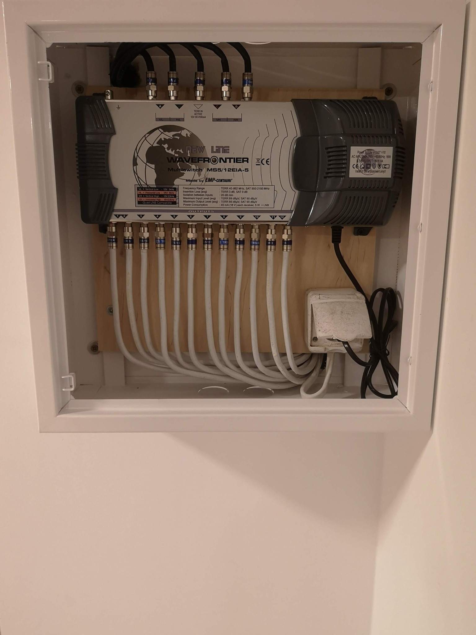 Rozdzielacz antenowy Wavefrontier MS5/12EIA-5 zamontowany w białej szafce z widocznymi kablami koncentrycznymi i zasilaczem, zainstalowany na drewnianej płycie wewnątrz szafki.