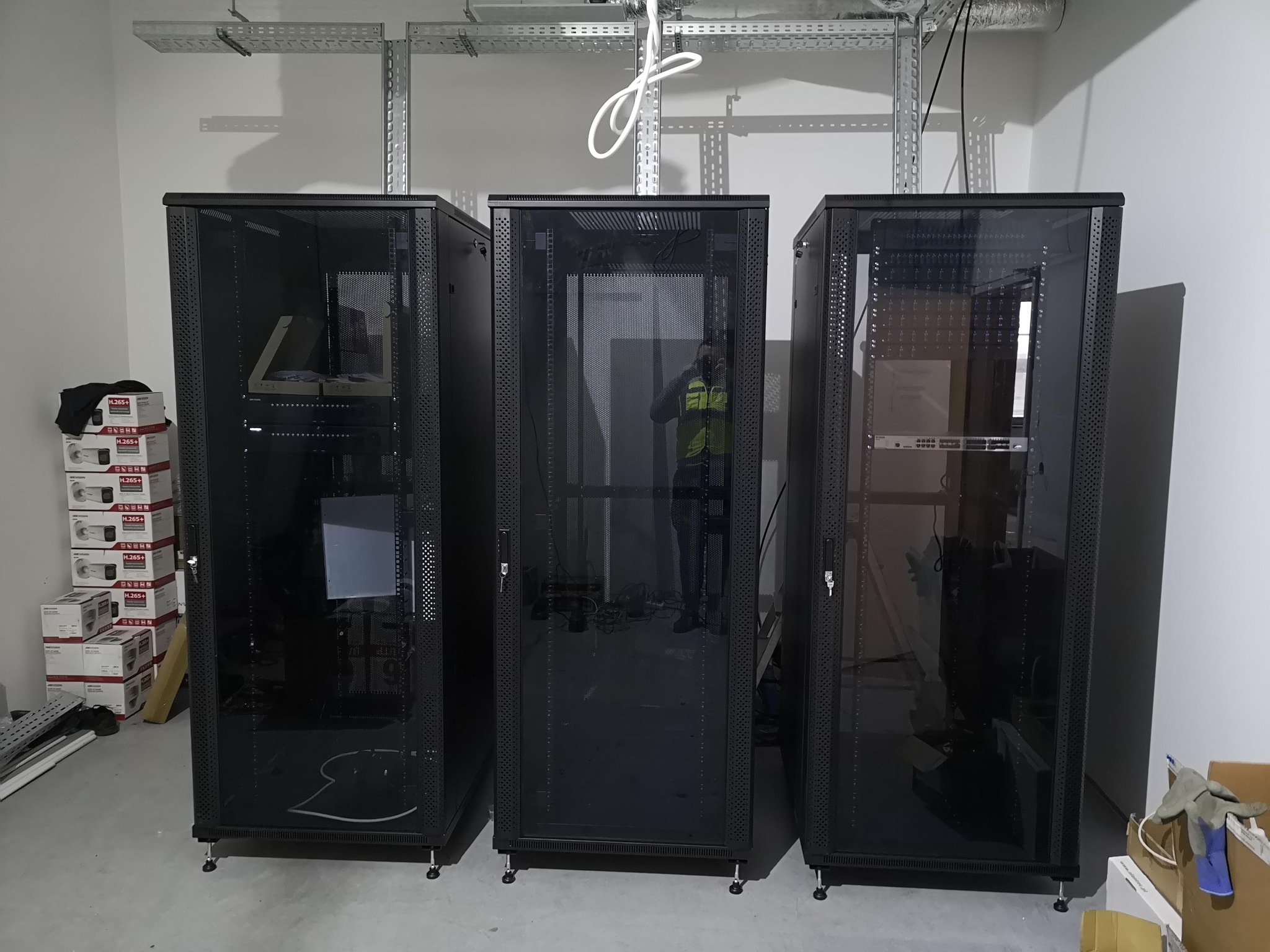 Trzy szafy serwerowe rack 19 cali w nowym pomieszczeniu, widoczne kable na suficie oraz pudełka z kamerami monitoringu na podłodze.