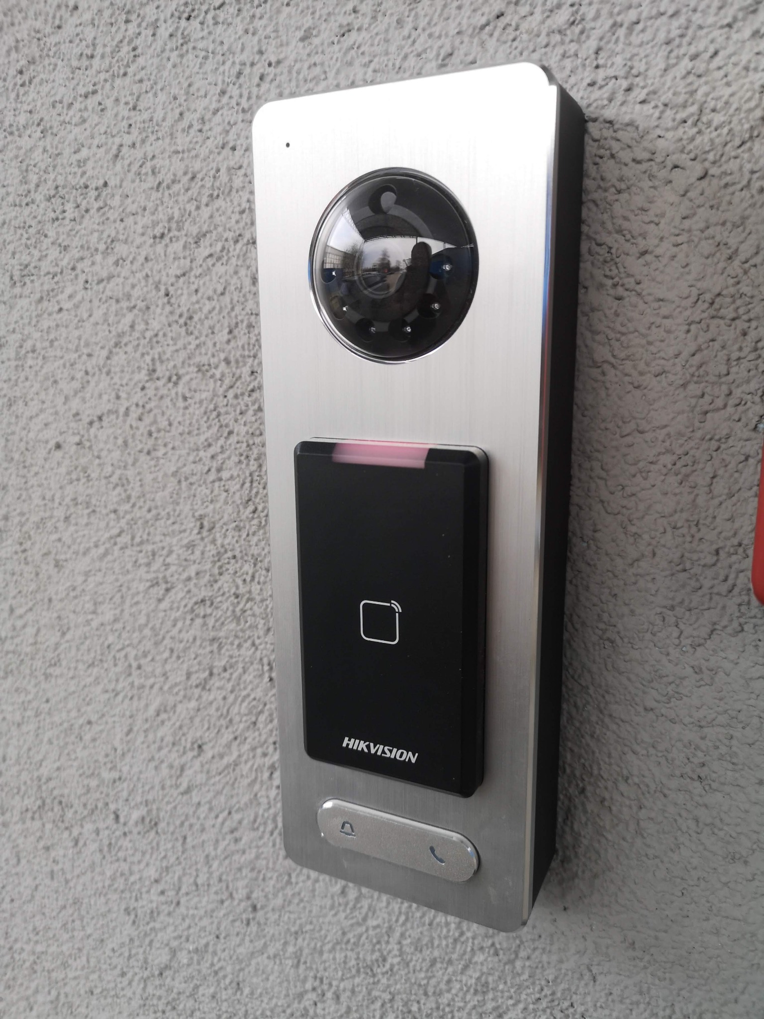 Zewnętrzny panel wideodomofonu Hikvision z kamerą, czytnikiem kart zbliżeniowych i przyciskiem dzwonka, zamontowany na szarej, chropowatej ścianie.