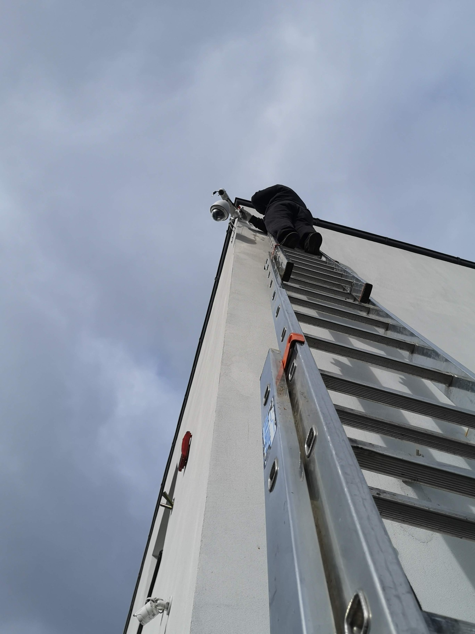Instalator na drabinie montuje kamerę monitoringu na elewacji budynku, widok z dołu w pochmurny dzień.