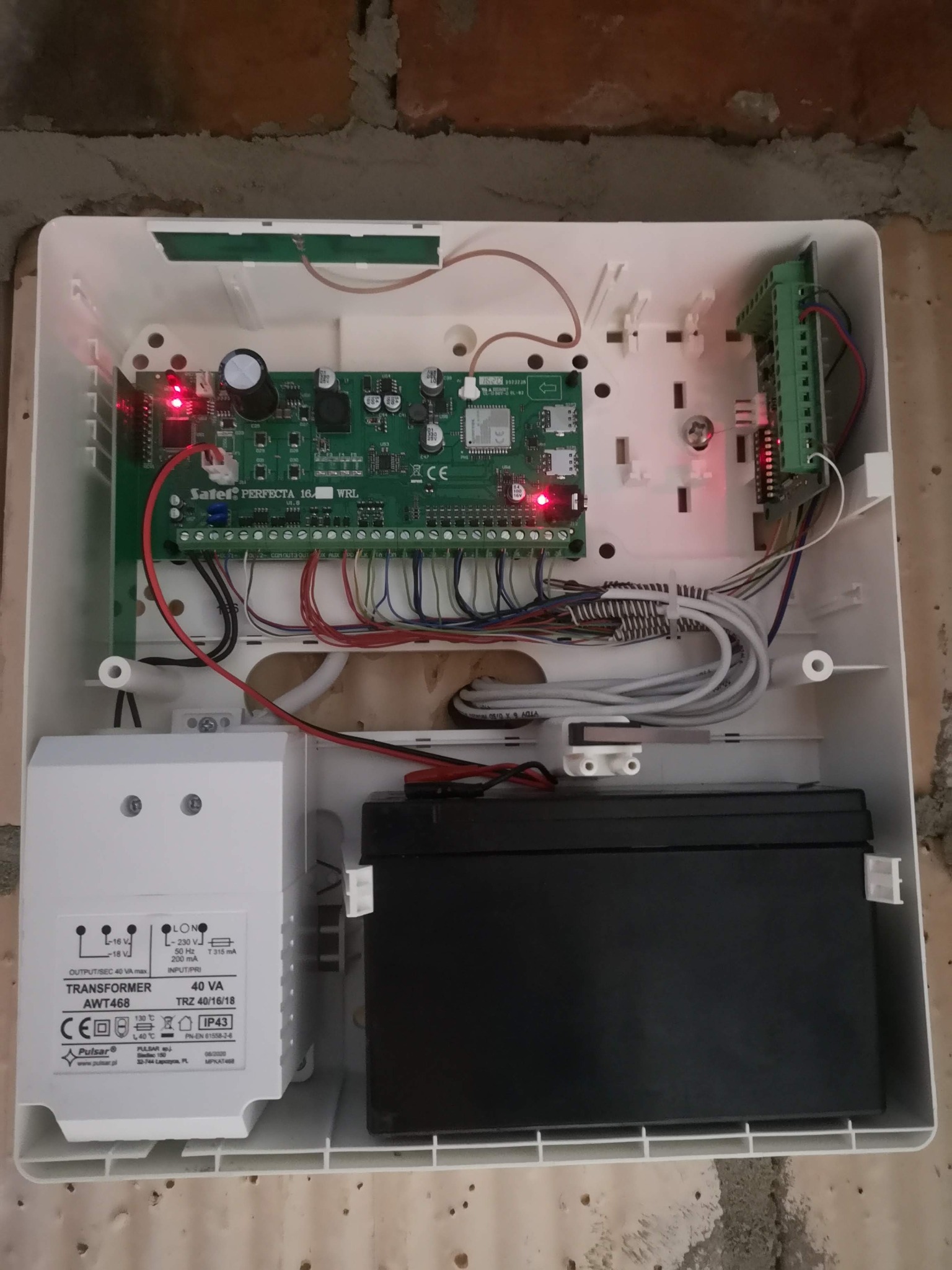 Otwarta obudowa centrali alarmowej Safetel Perfecta 16 WRL z widoczną elektroniką, transformatorem Pulsar AWT468 i akumulatorem, zamontowana na ścianie z cegły.