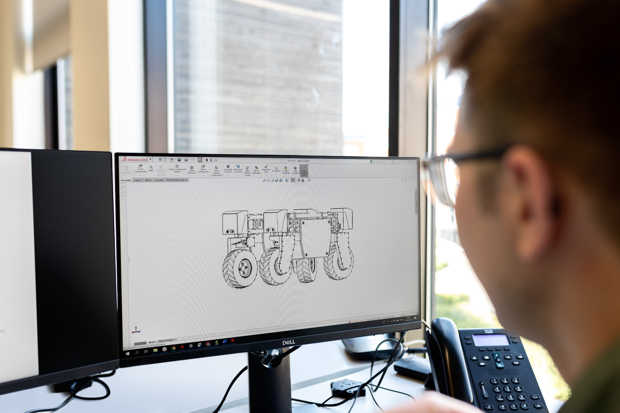 Inżynier w okularach pracuje nad projektem ciągnika rolniczego w programie SolidWorks na podwójnym monitorze, widoczne okno z rysunkiem technicznym 3D.